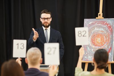 An auctioneer taking bids from the rostrum as numbered paddles are raised in front of a painting